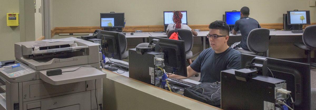 Students with laptops sit around a table and collaborate on a project while look at two large monitors