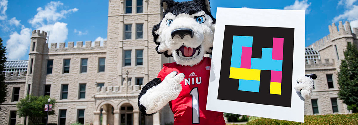 NIU's mascot, Victor E. Huskie, holds a NaviLens QR code saying the building has NaviLens wayfinding installed.