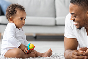 happy African American baby sits up on soft rug and plays with father