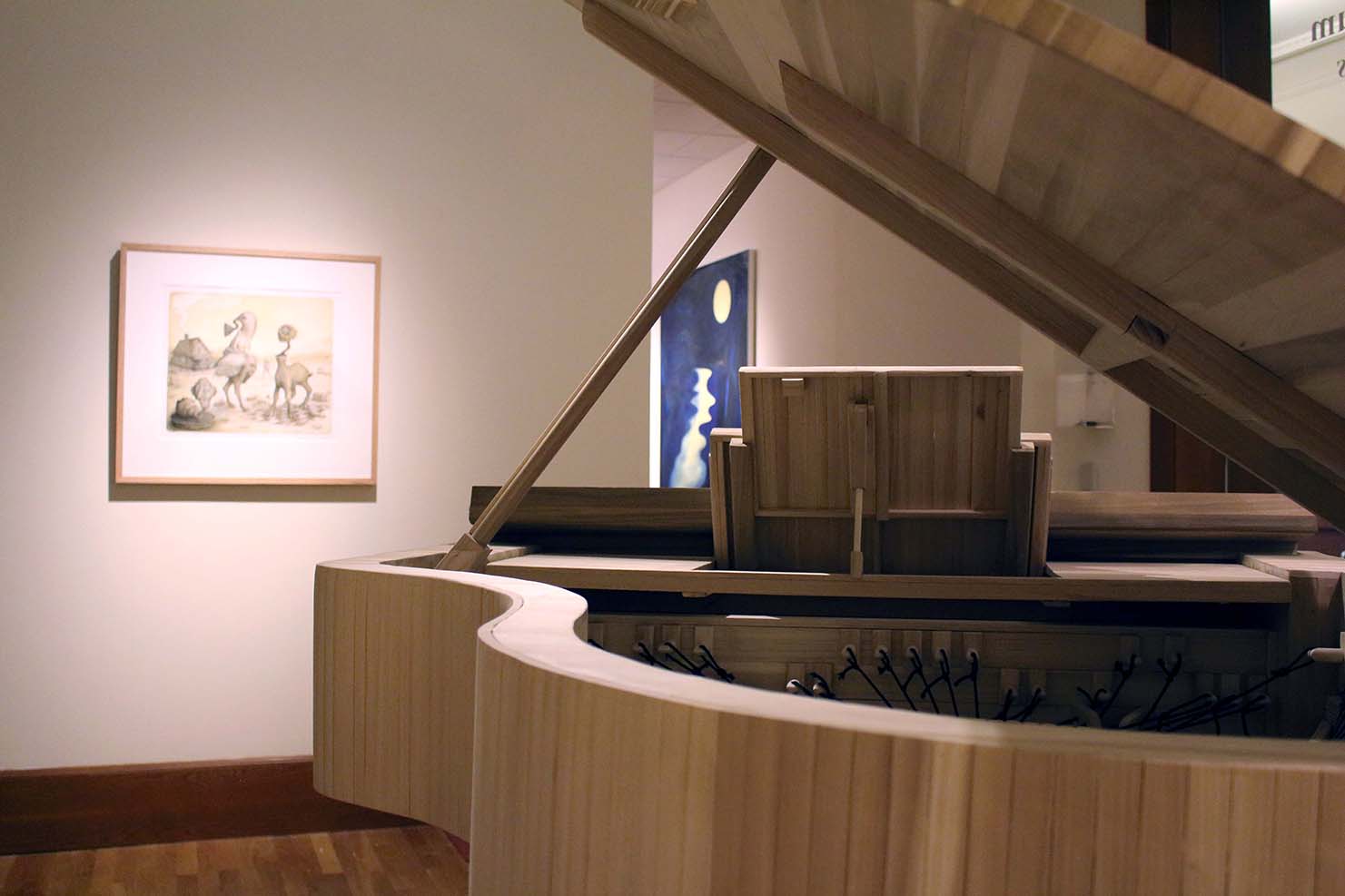 Installation image of grand constructed wood piano sculpture and lithographic print in background.