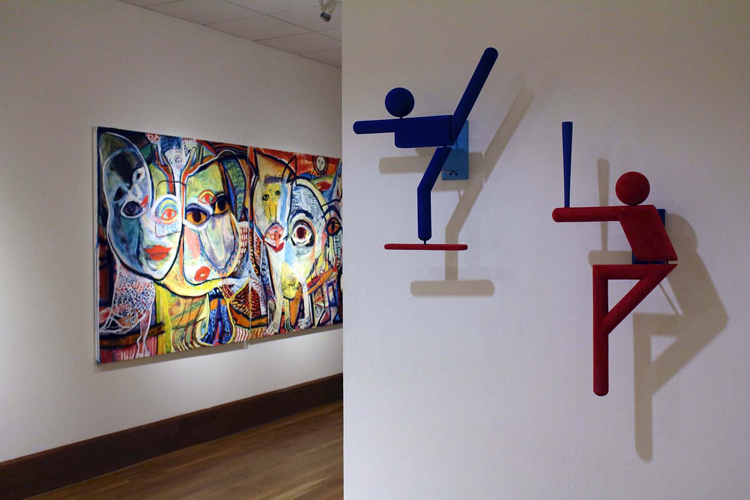 Foreground installation view of blue and red universal graphic sculptures of karate and baseball figures with expressionistic colorful oil on canvas painting in background.