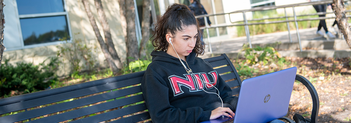 woman wearing an NIU hooded sweatshirt student sitting on a metal bench outside, using a laptop and wearing earbuds