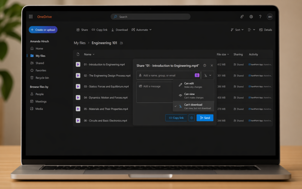 A laptop sits on a wooden desk displaying a OneDrive interface in dark mode. The page shows a folder titled “Engineering 101” under “My files,” with a list of video lecture files for an introductory engineering course, along with file sizes and sharing status. On the left sidebar are navigation options such as “Home,” “My files,” “Shared,” “Favorites,” and “Recycle bin,” followed by a “Browse files by” section. Centered on the screen is a sharing pop-up window for one video file, with fields to add a name or email, an optional message, and permission settings. The “Can’t download” option is selected, and buttons labeled “Copy link” and “Send” appear at the bottom.