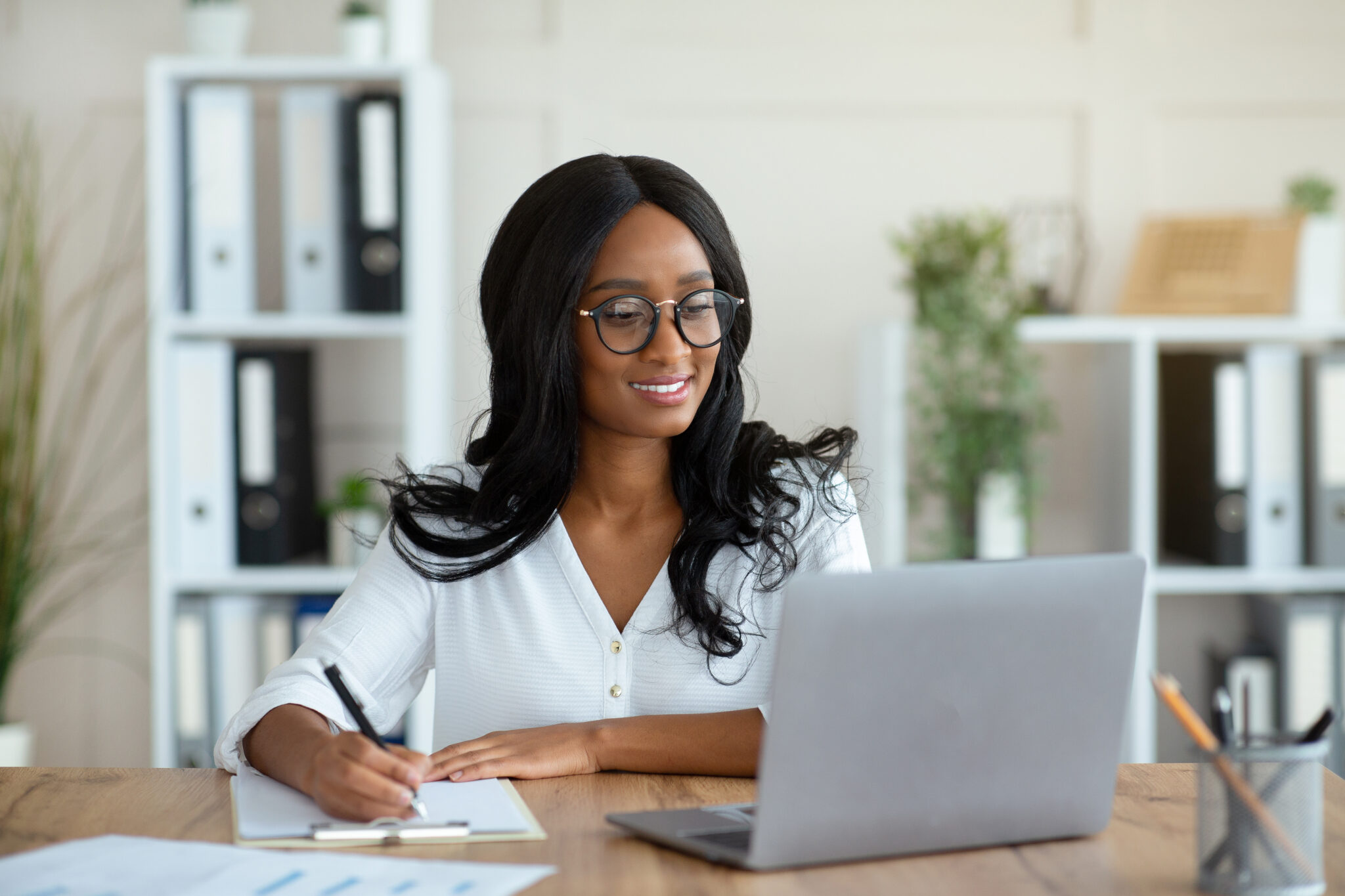 woman sitting at a desk smiling while looking at laptop computer and writing on paper beside laptop