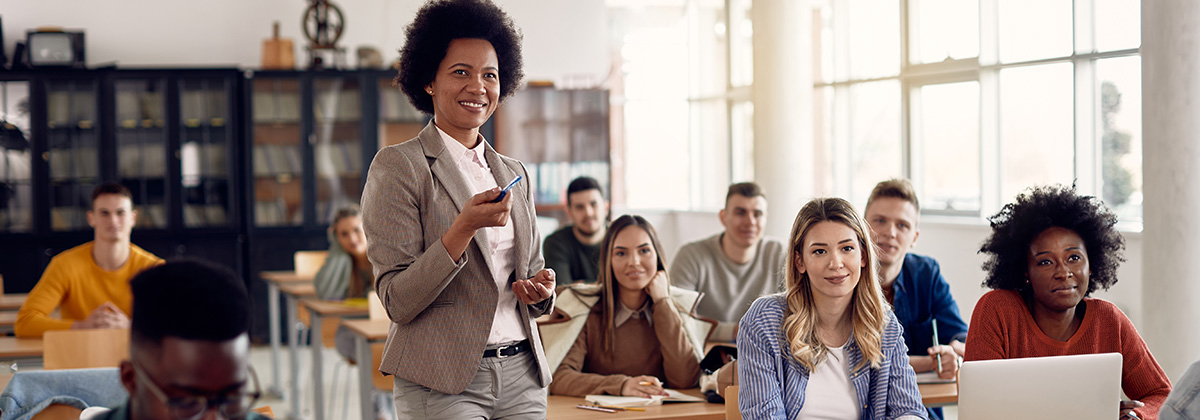 Black woman professor confidently gesturing to the front of the room in a classroom with multiple students at desks