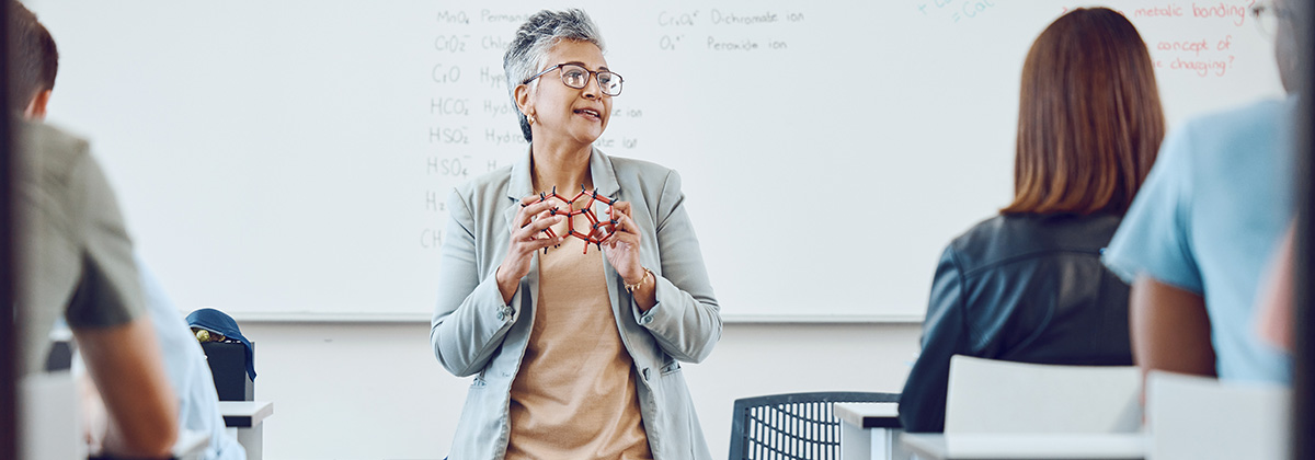 Female professor with short gray hair leans against a desk and holds a molecular model while presenting to students sitting in desks