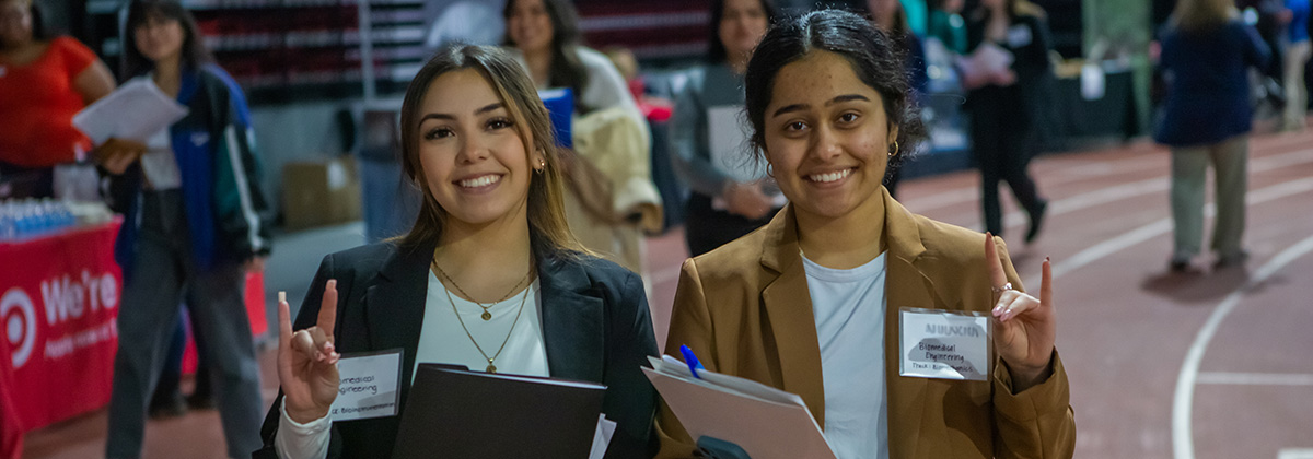 two female students wearing blazers make a huskie sign with their hands, background is the NIU career fair