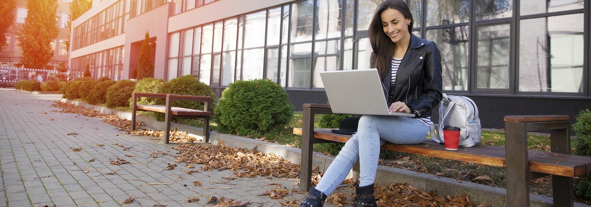 woman sitting on a bench in front of a university building, using a laptop