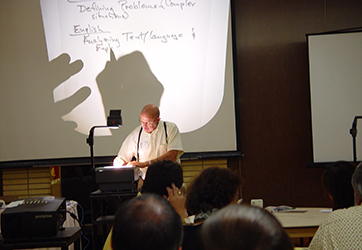 man writes on overhead projector transparency while presenting to a room of people
