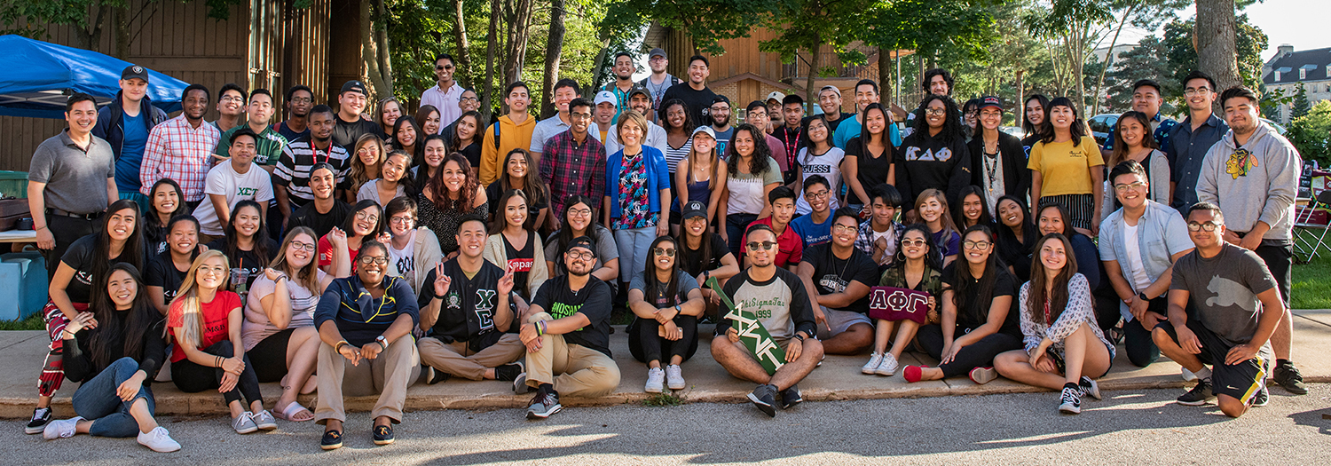 Large group of diverse college students and staff smiling and posing together outdoors on a sunny day, seated and standing in rows near a wooden pavilion and trees for the Asian American Resource Center