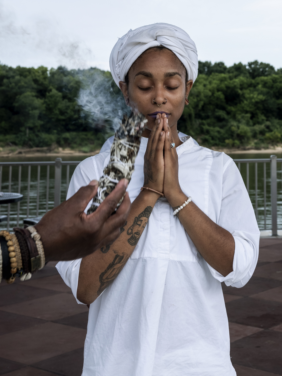 woman in white praying as burning sage is held before them by a river.