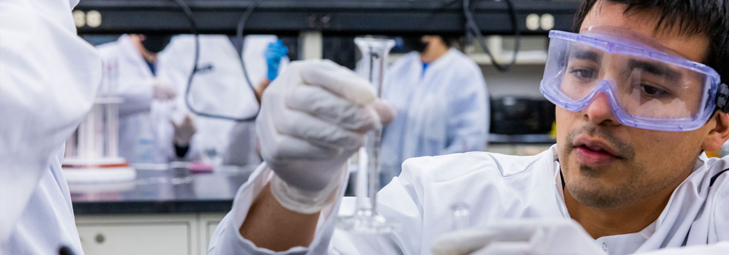 Male Mac lab student with safety glasses, looking at slim,  glass beaker.