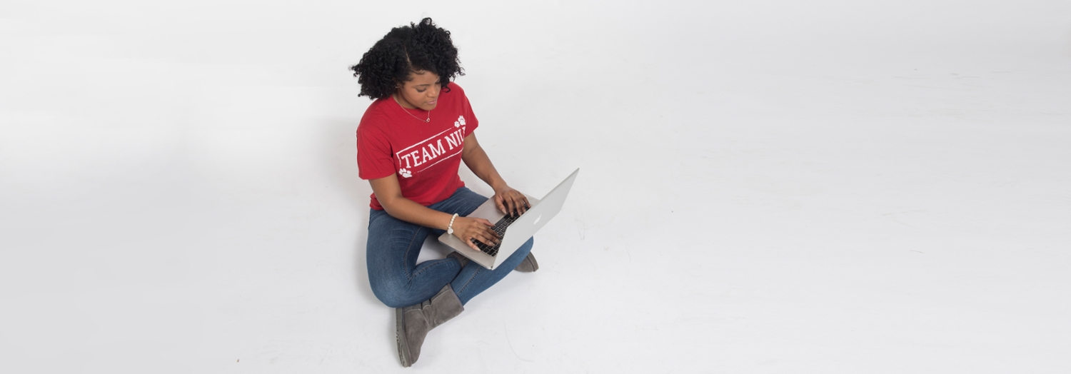 Student in NIU tshirt sitting on floor with laptop