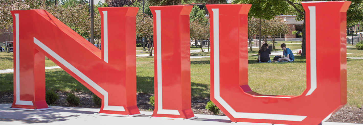 NIU lettering monument with students sitting on grass in background