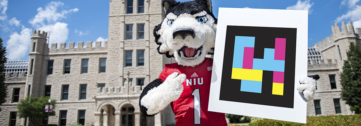 NIU's mascot, Victor E. Huskie, holds a NaviLens QR code saying the building has NaviLens wayfinding installed.