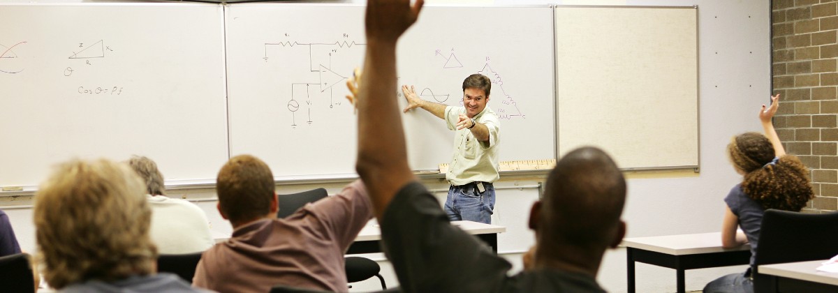 professor writing on dry erase marker board, students in foreground with hands raised