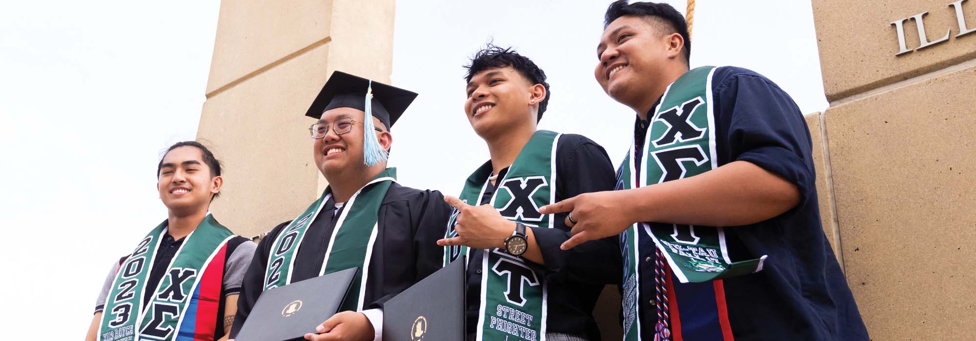 Members of Chi Sigma Tau by the bell outside of the Convocation Center after a commencement ceremony dressed in graduation clothing