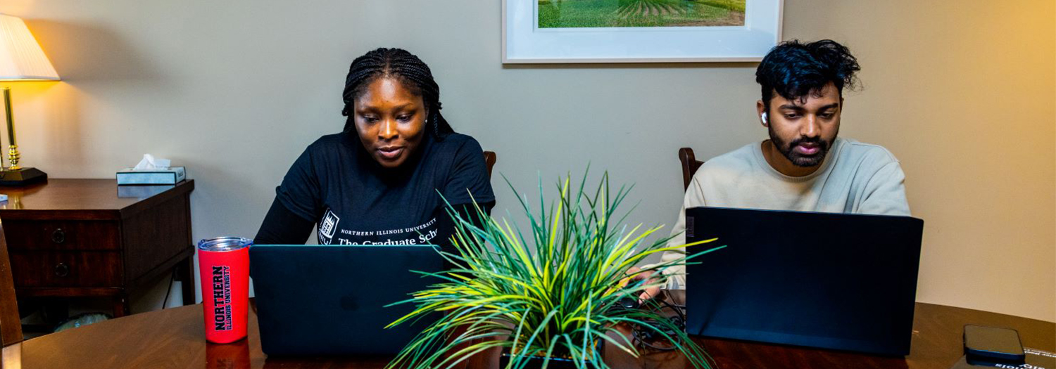 Two students work on laptops at a table with a plant in the center.