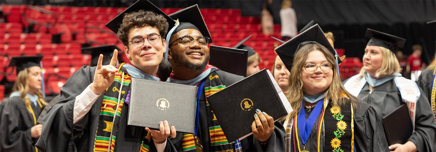 Three graduates wearing caps and gowns smile while holding their diplomas during a commencement ceremony, standing together in a crowded indoor arena