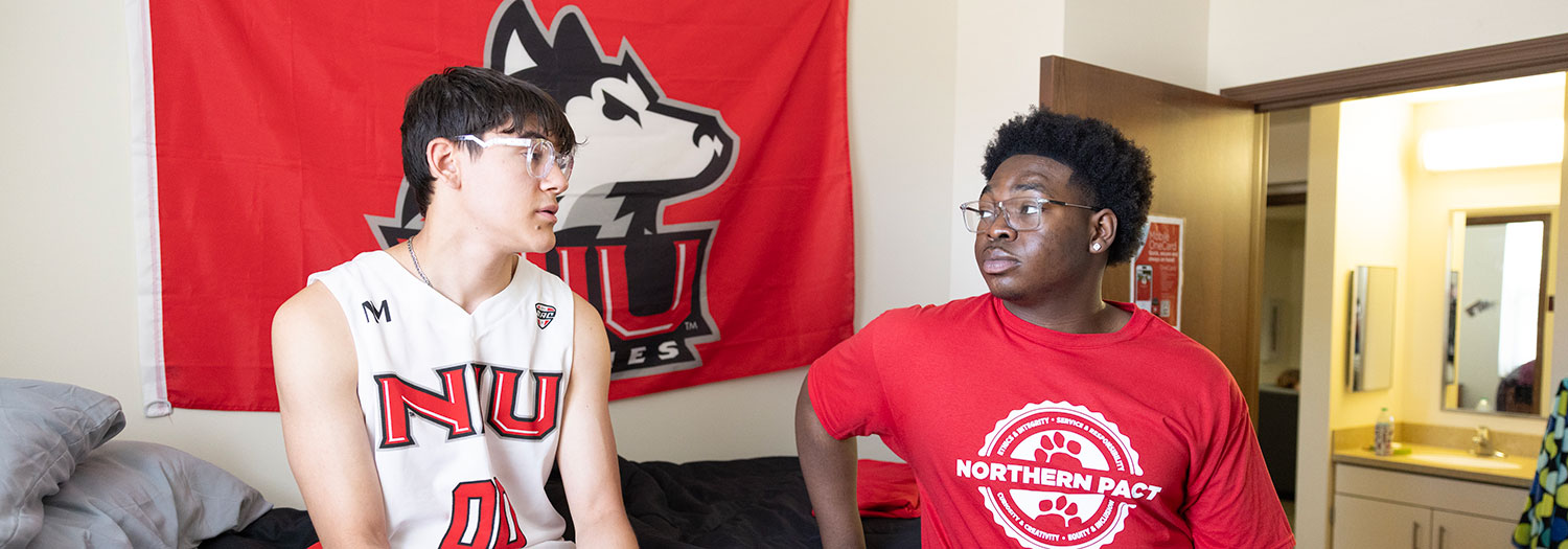 Two residents talking in their room with an NIU Huskies flag in the background.