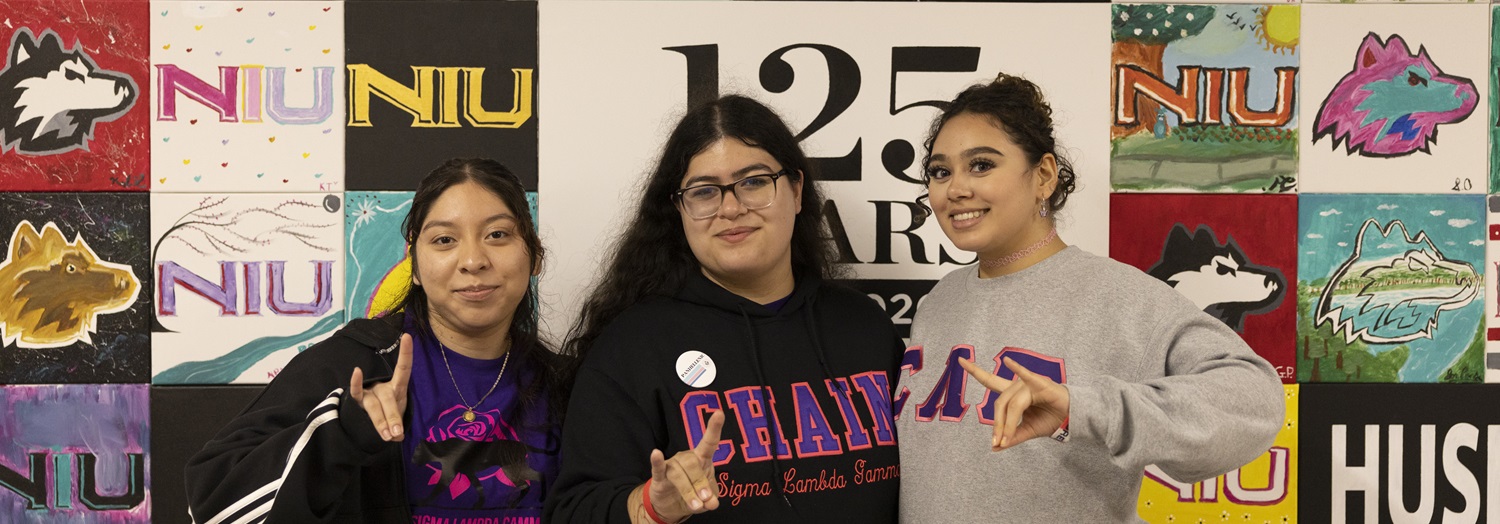 Three students pose in front of an NIU mural at the Holmes Student Center