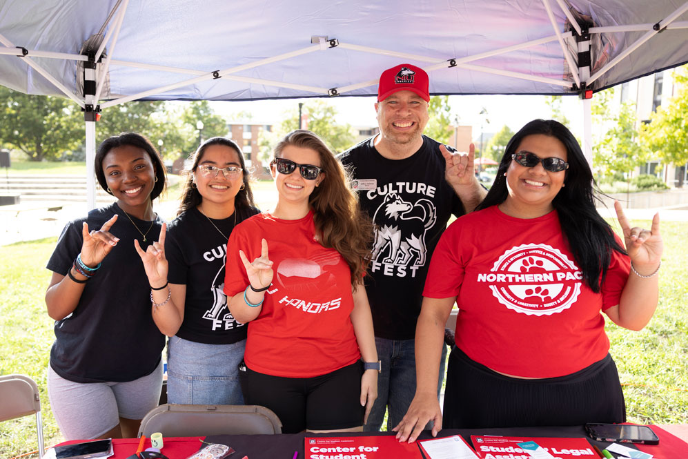 Five students and a staff member at a Culture Fest table on campus, making the Huskie hand sign