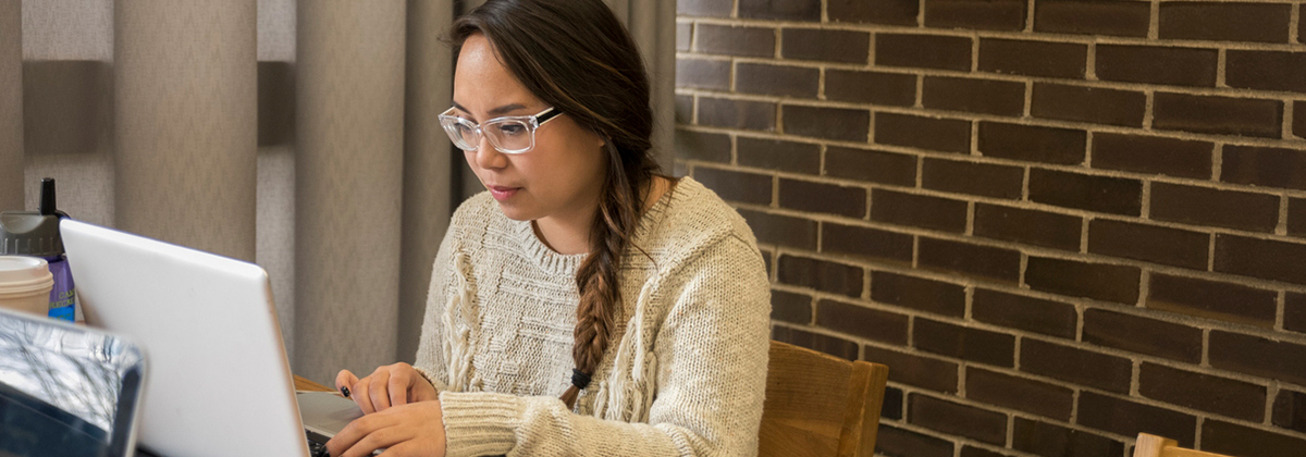 A student typing on a laptop in front of a brick wall in Founders Memorial Library